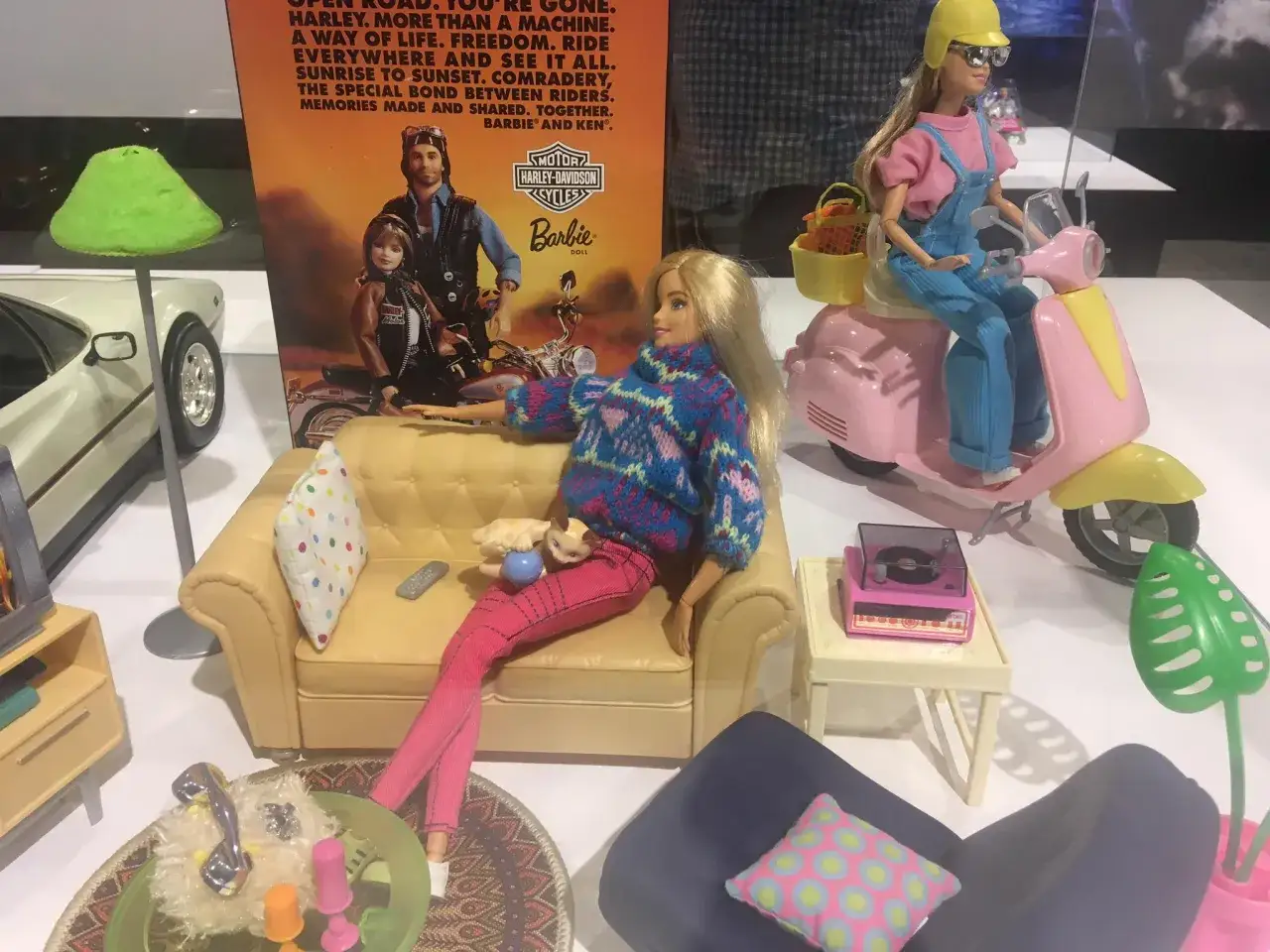 Wystawa muzeum sztuk użytkowych Barbie: lalka na kanapie, druga na skuterze, a w tle plakat z Kenem i Barbie na motocyklach Harley-Davidson.