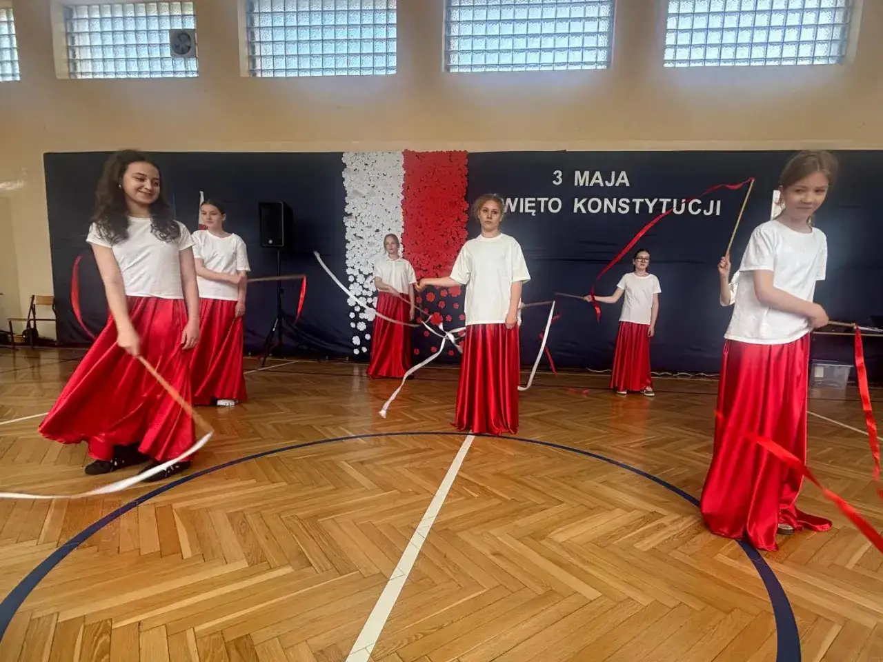 Dzieci w białych koszulkach i czerwonych spódnicach tańczą z wstążkami, celebrując Święto Konstytucji 3 Maja.