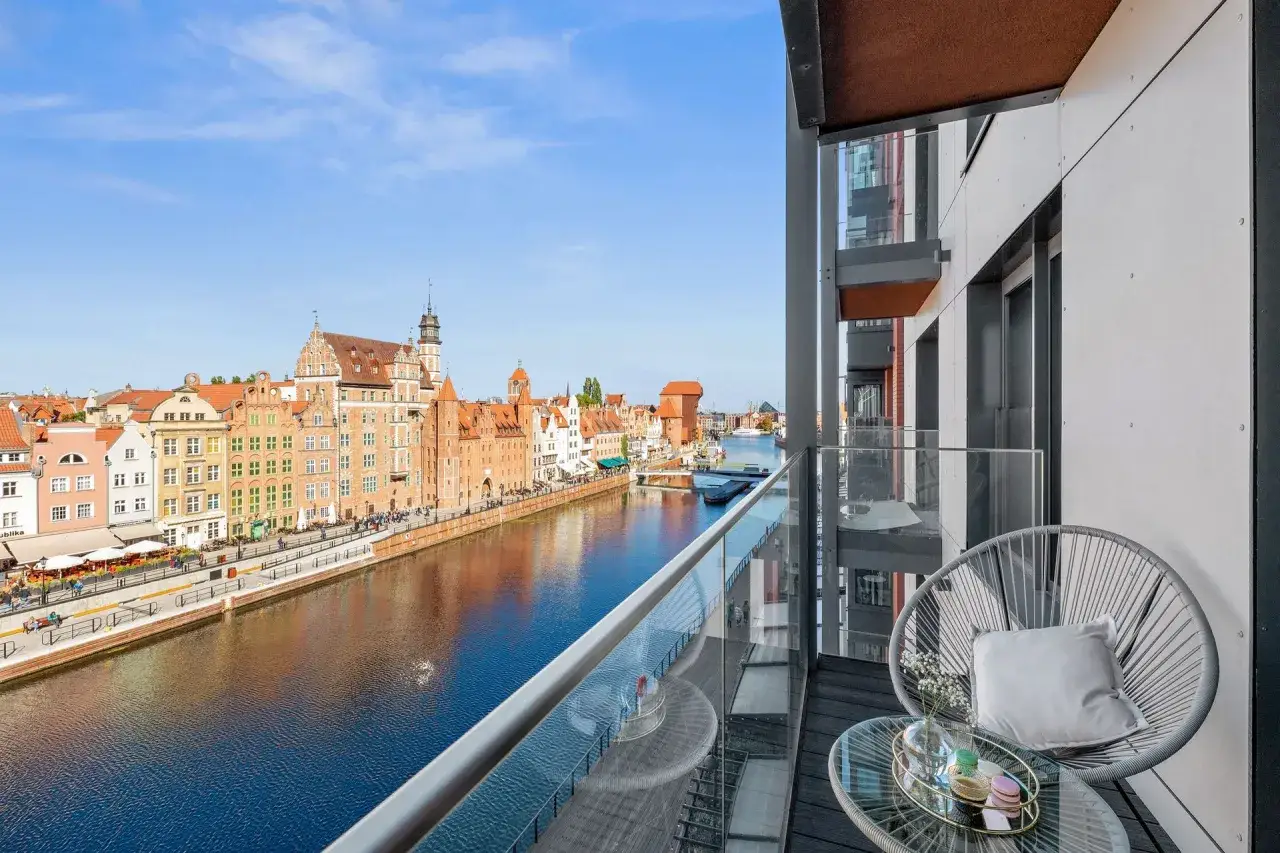 Widok z balkonu na Motławę i zabytkowe kamienice w Gdańsku. Na pierwszym planie nowoczesny balkon z fotelem.