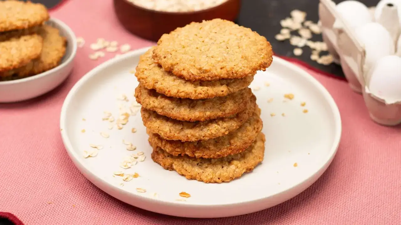 Ein Stapel goldbrauner Haferkekse, perfekt für ein schnelles Cookies Rezept. Zutaten wie Haferflocken und Eier sind im Hintergrund zu sehen.