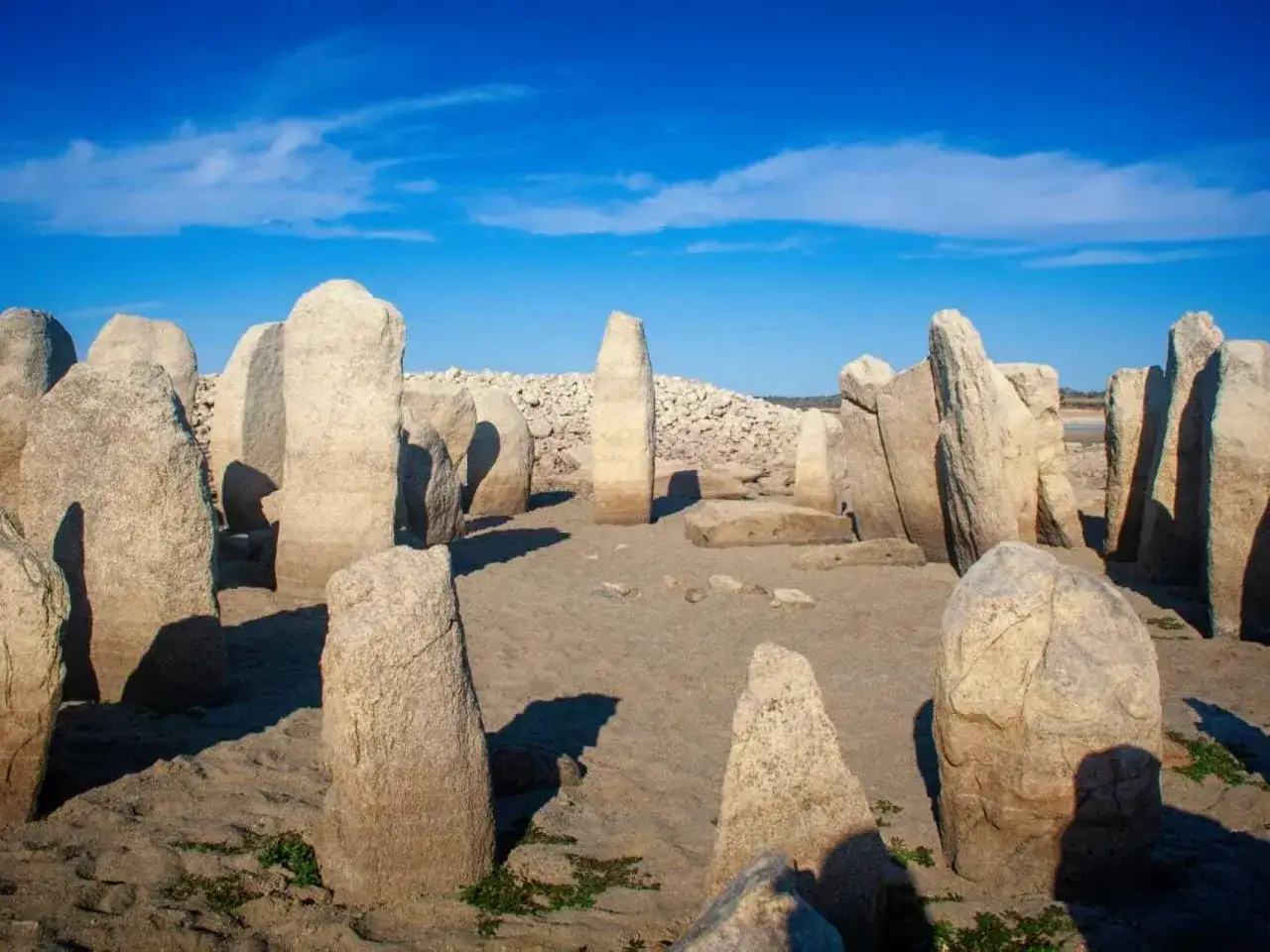 Un círculo de grandes piedras verticales, que es un cromlech, se alza sobre un terreno arenoso bajo un cielo azul brillante.