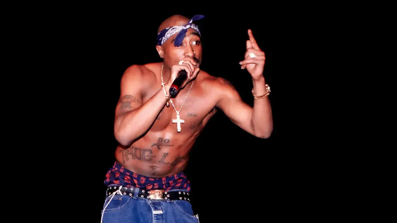 Tupac Shakur na scenie, z mikrofonem w dłoni, w bandanie i z tatuażem