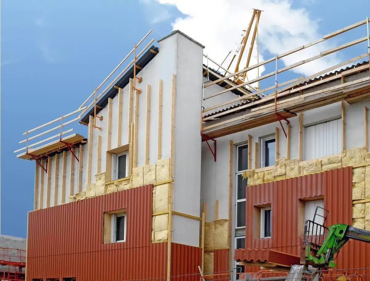 Chantier de ravalement avec isolation ext&eacute;rieure en cours. Des panneaux de bardage rouge et de la laine de roche sont visibles.