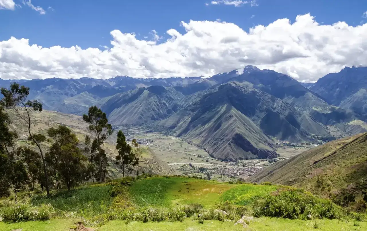 Grüne Terrassenfelder und Bäume blicken auf ein Dorf im Tal der **Peru Anden**. Majestätische Berge erheben sich im Hintergrund unter einem blauen Himmel mit weißen Wolken.