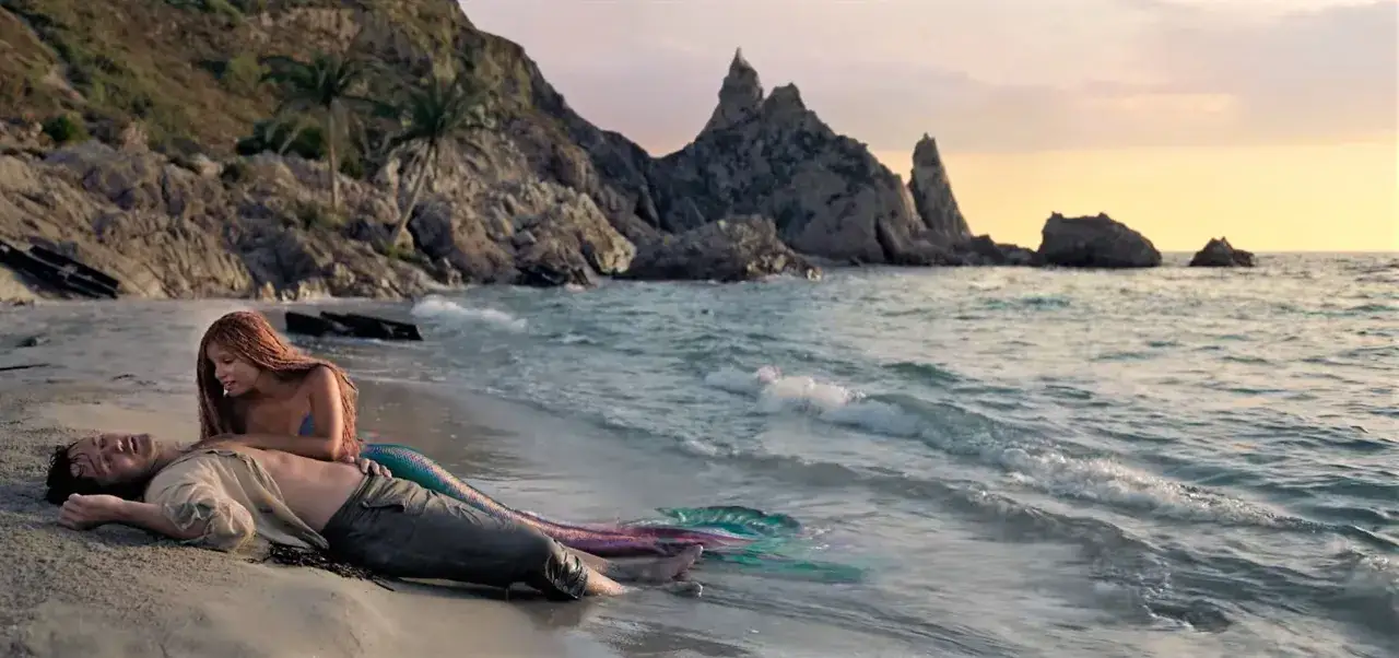 Una sirena con la coda colorata si china su un uomo naufrago sulla spiaggia. Travolti da un insolito destino, la curiosità nei suoi occhi è palpabile.