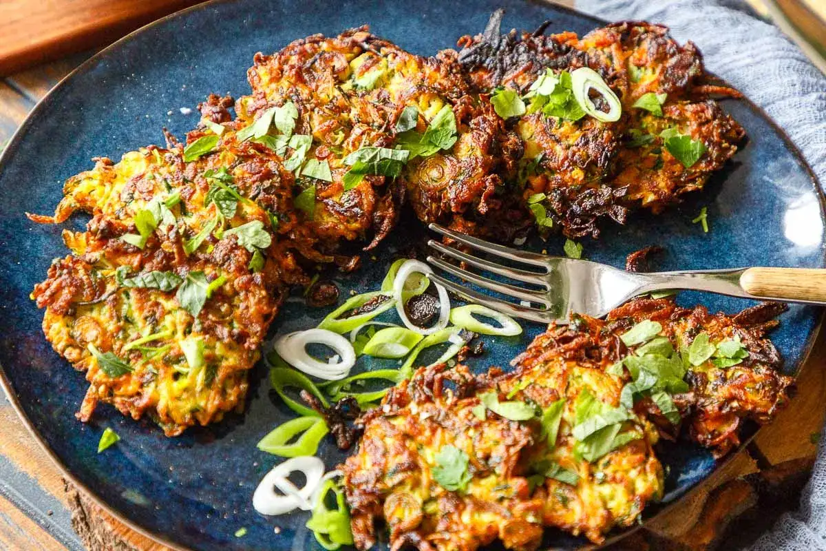 Knusprige Zucchini-Puffer, garniert mit frischen Kr&auml;utern und Fr&uuml;hlingszwiebeln, liegen auf einem blauen Teller. Eine Gabel liegt daneben.