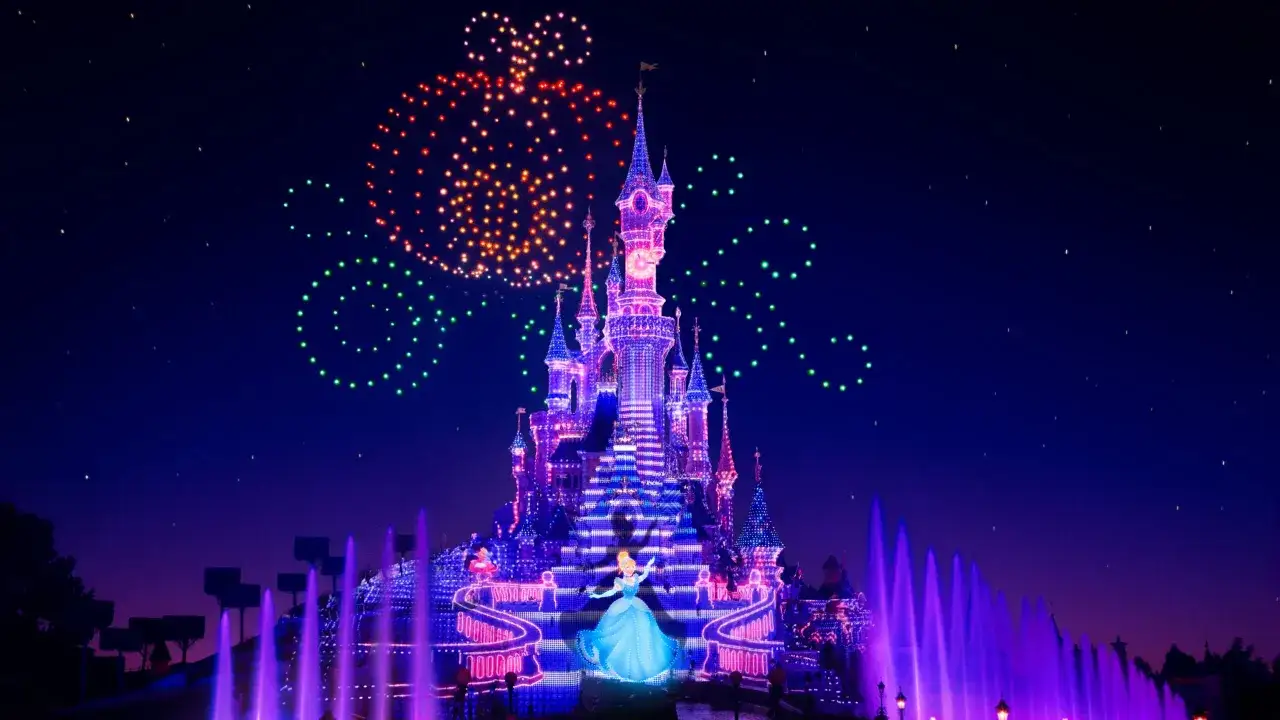 Castillo iluminado en Disneyland Paris con fuegos artificiales en forma de Mickey y fuentes moradas.