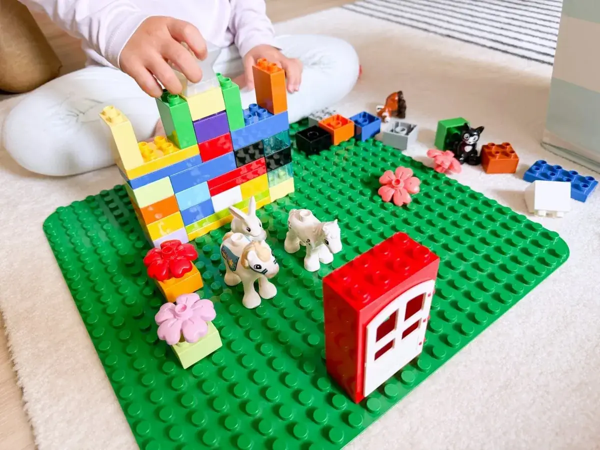 Kind baut mit bunten Duplo Steinen eine Burg. Viele Duplo bauen Ideen sind auf der gr&uuml;nen Platte zu sehen, darunter Tiere und Blumen.