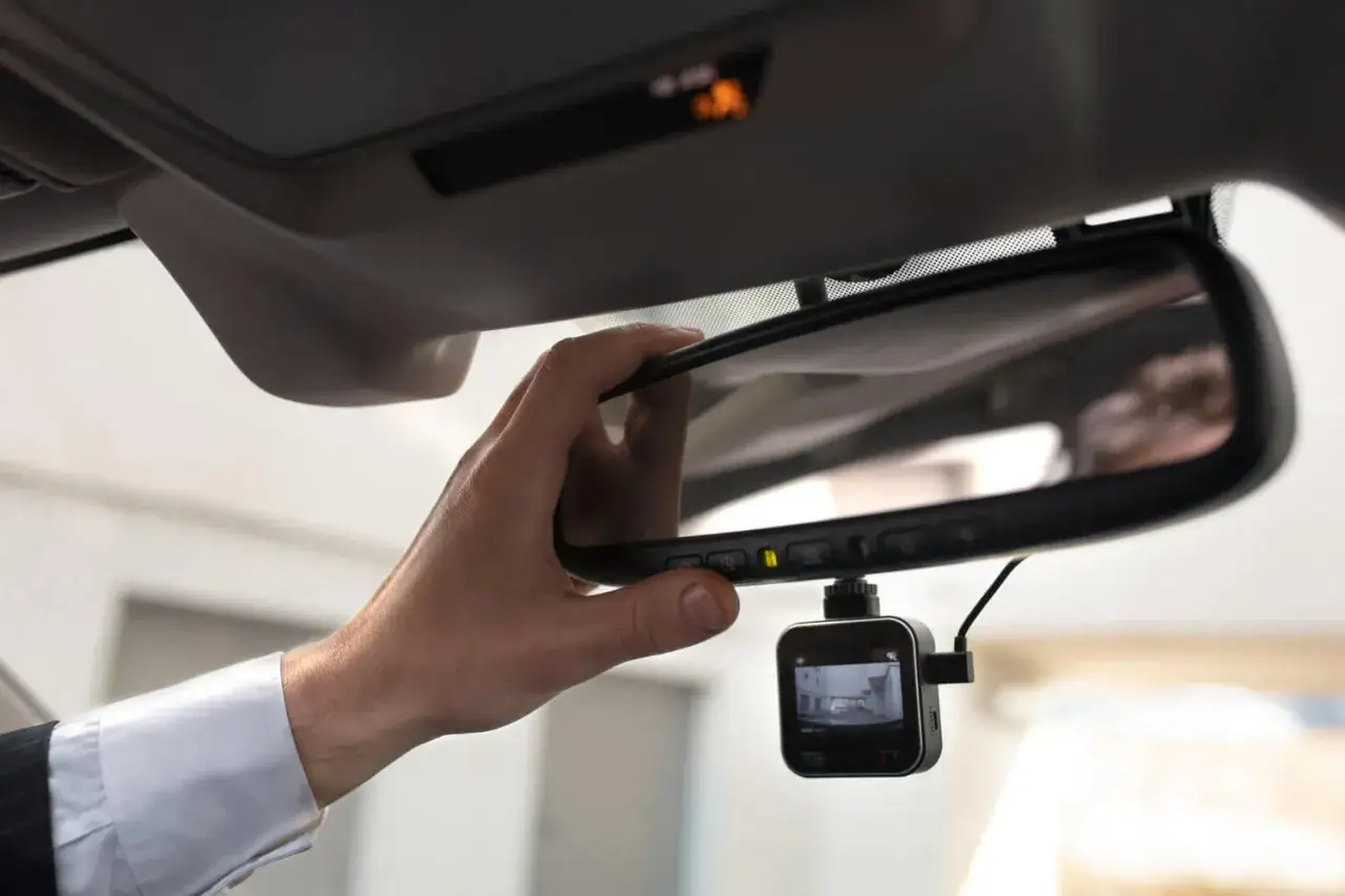 Mano che regola lo specchietto retrovisore, sotto cui è agganciata una telecamera per auto antivandalismo, legale e discreta.