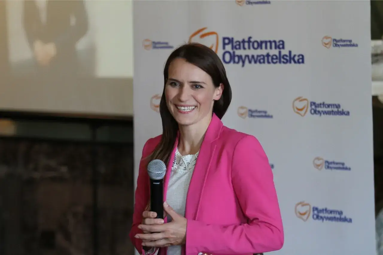 Agnieszka Pomaska, z uśmiechem i mikrofonem w dłoni, na tle logo Platformy Obywatelskiej. Jej pochodzenie jest związane z polityką.