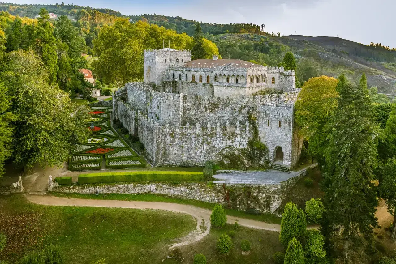 El castillo de Pambre, una fortaleza de piedra rodeada de exuberante vegetación y jardines formales, se alza majestuoso en un paisaje montañoso.