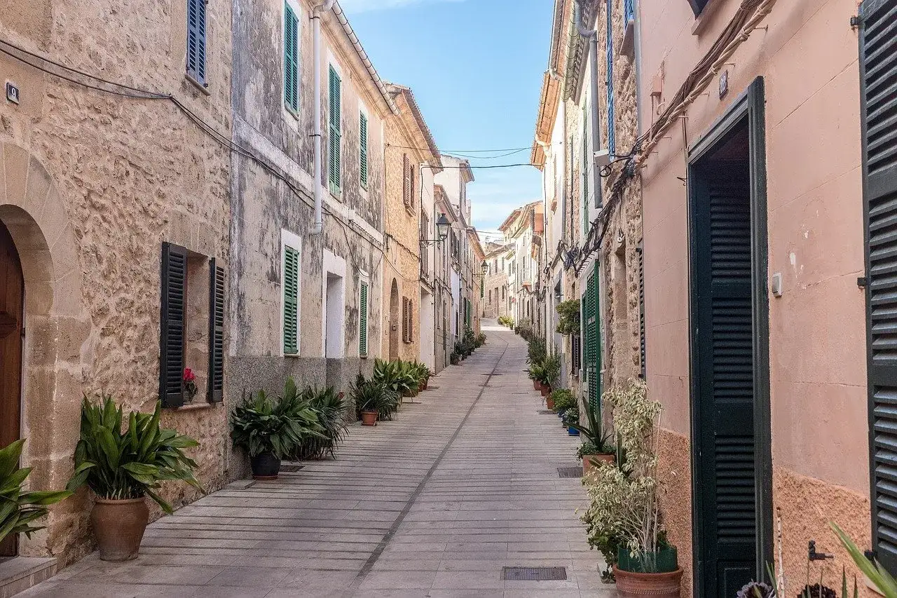 En smal, stenlagd gata i Alcudia old town kantas av gamla hus med gr&ouml;na f&ouml;nsterluckor och krukv&auml;xter.
