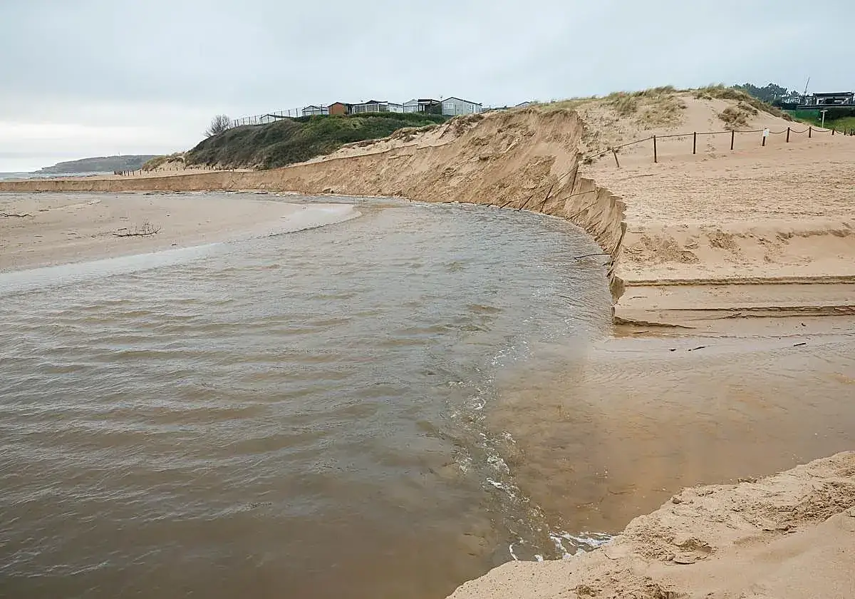 Desembocadura de un río en la playa de Loredo, Cantabria, con dunas erosionadas y casas al fondo.