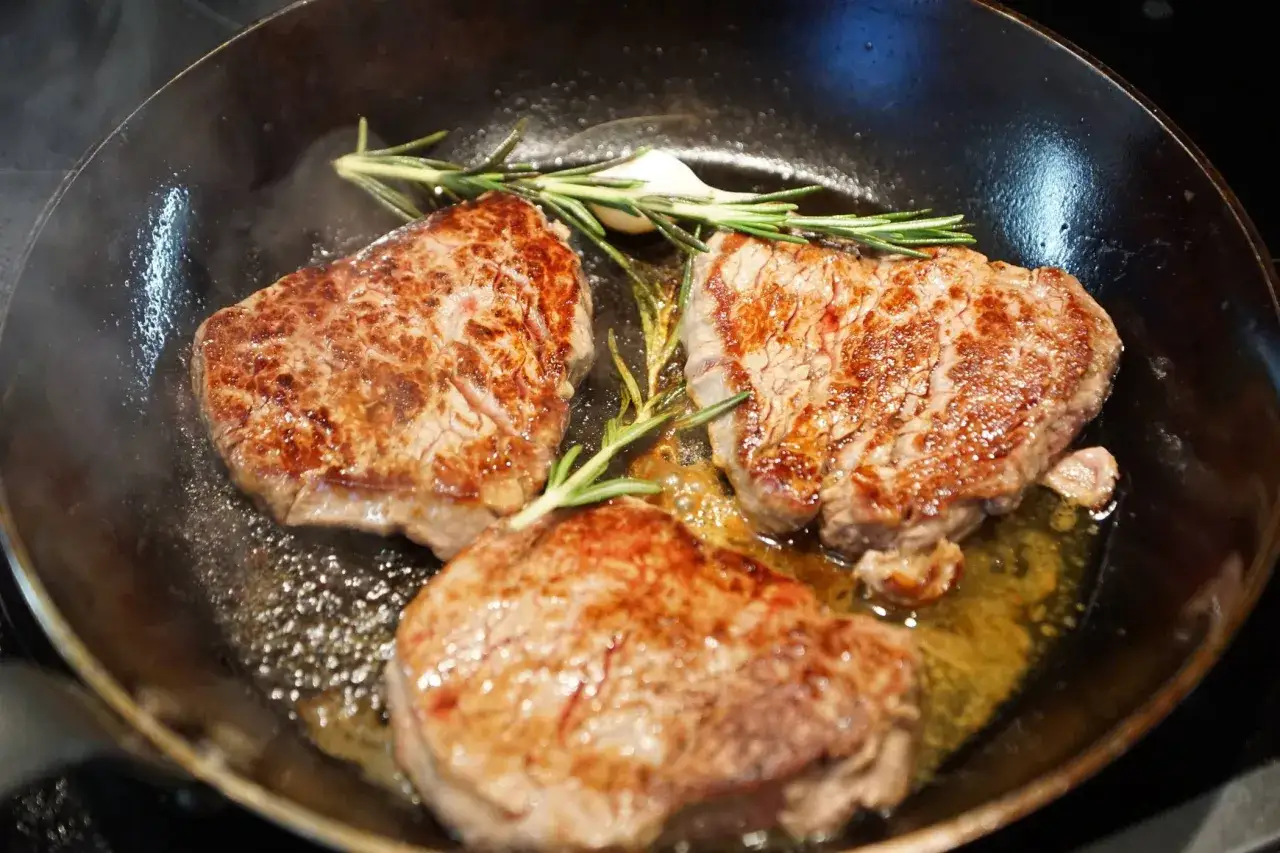 Drei saftige Steaks braten in einer Gusseisenpfanne mit Rosmarin und Knoblauch.