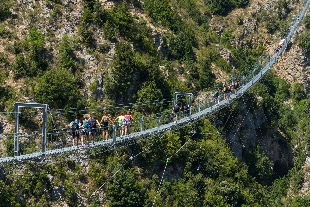 Persone camminano su un lungo percorso ponte tibetano sospeso tra montagne verdi.