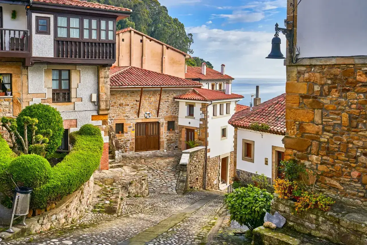 Calle empedrada en uno de los pueblos con encanto en Asturias. Casas de piedra y tejados rojos descienden hacia el mar azul.