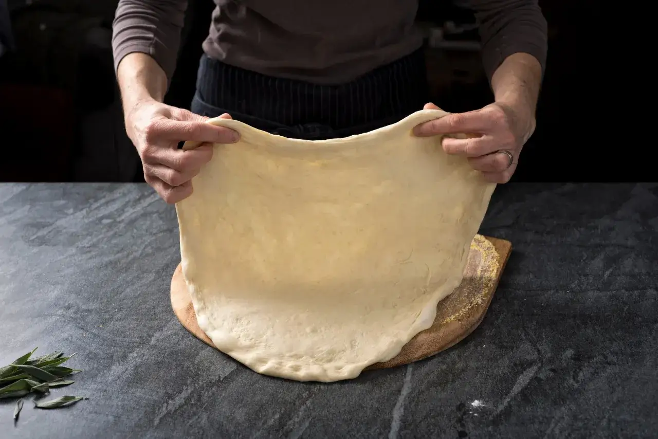 Des mains étirent une pâte à pizza souple, prête pour une recette rapide. Une touche de farine et des herbes apportent fraîcheur.