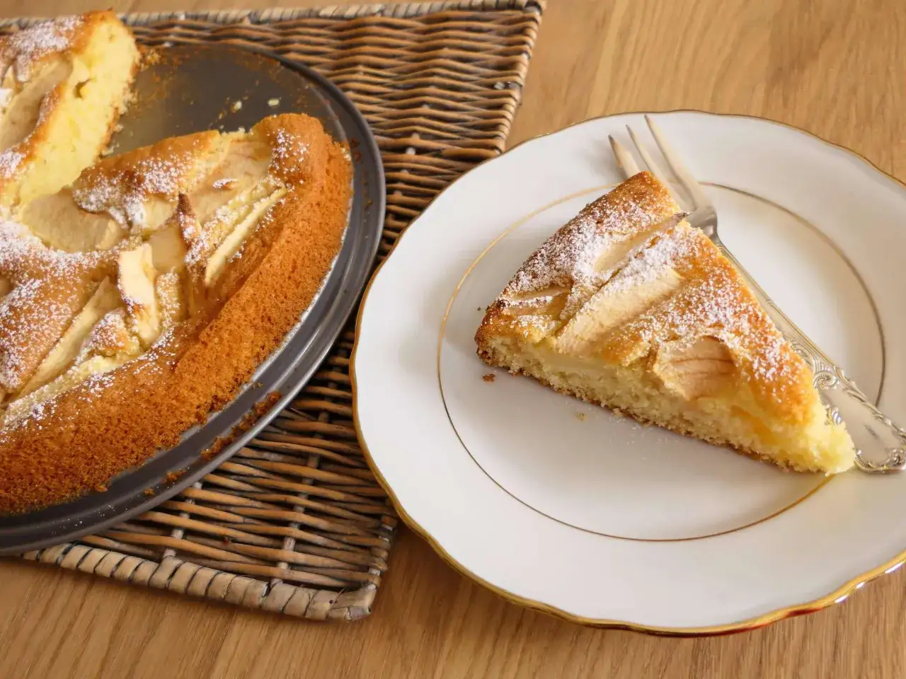 Ein Stück apfelkuchen sehr fein, mit Puderzucker bestäubt, liegt auf einem Teller. Daneben ist der Rest des Kuchens in einer Form.