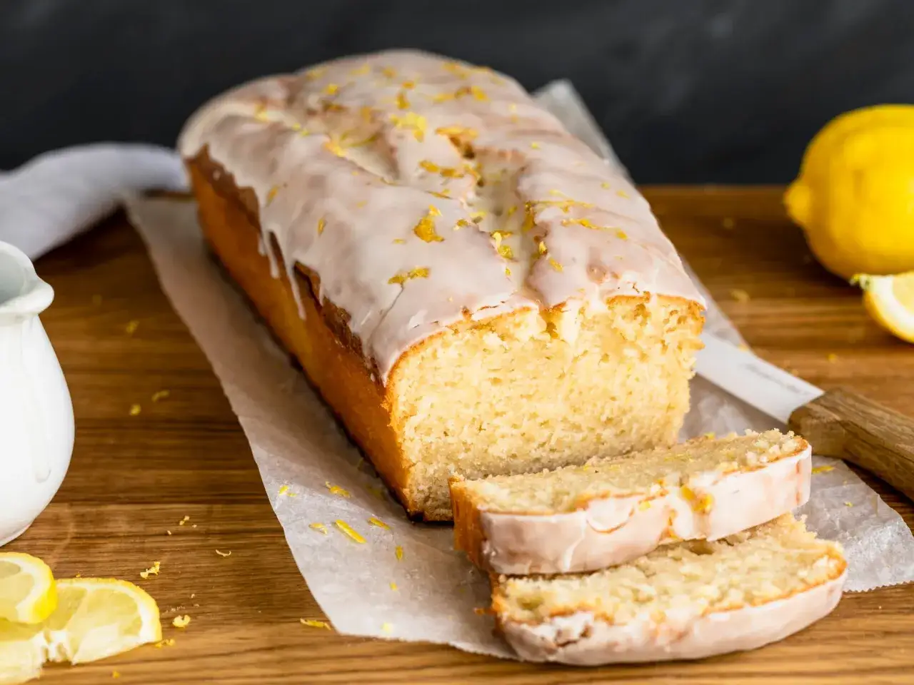 Saftiger veganer Zitronenkuchen mit Zuckerguss und Zitronenschale bestreut, daneben frische Zitronen.