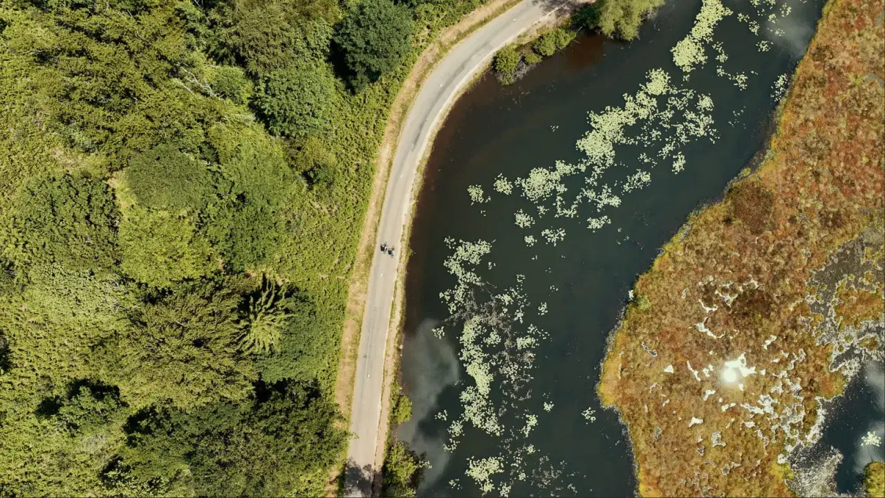 Vue aérienne d'un étang de 1000m2 bordé de végétation luxuriante. Une route sinueuse longe l'eau où flottent des nénuphars. Deux cyclistes pédalent paisiblement.