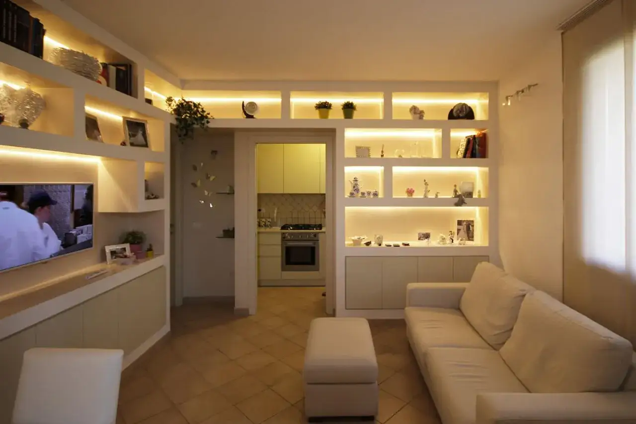 Illuminata libreria cartongesso con oggetti decorativi, foto e libri. Divano bianco e pouf in primo piano.