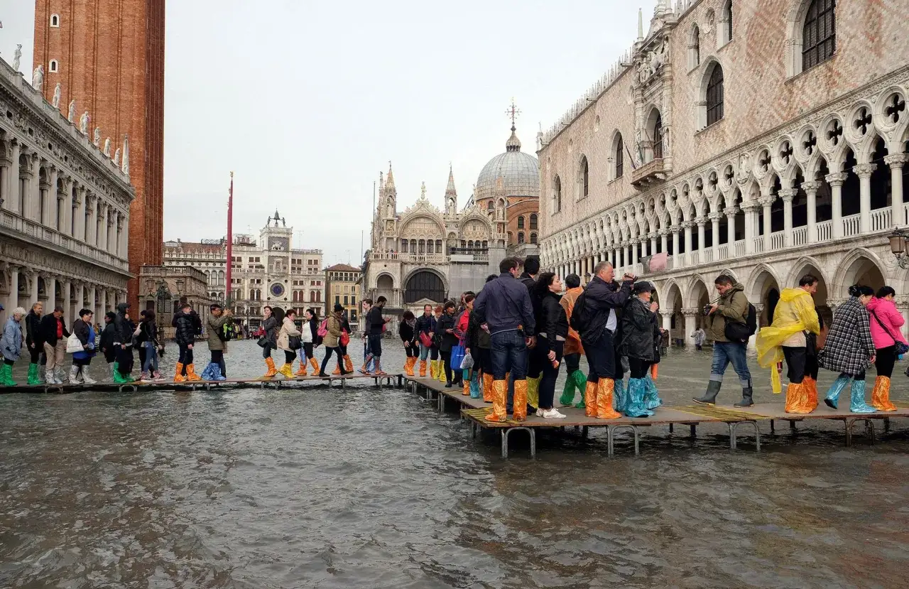 Gente con botas de agua camina sobre pasarelas en Venecia cuando se inunda. Edificios históricos y la Basílica de San Marcos al fondo.