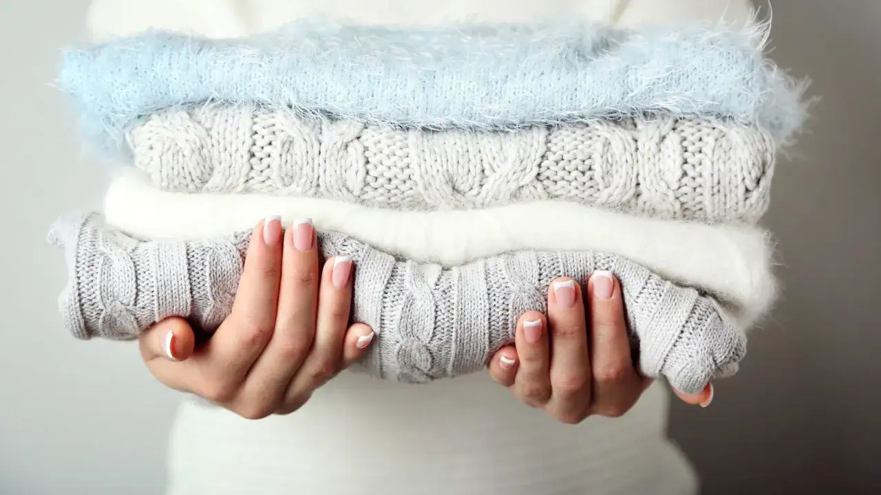 Mains soignées tenant une pile de pulls douillets. Idéal pour savoir comment detacher un pull blanc et le garder comme neuf.