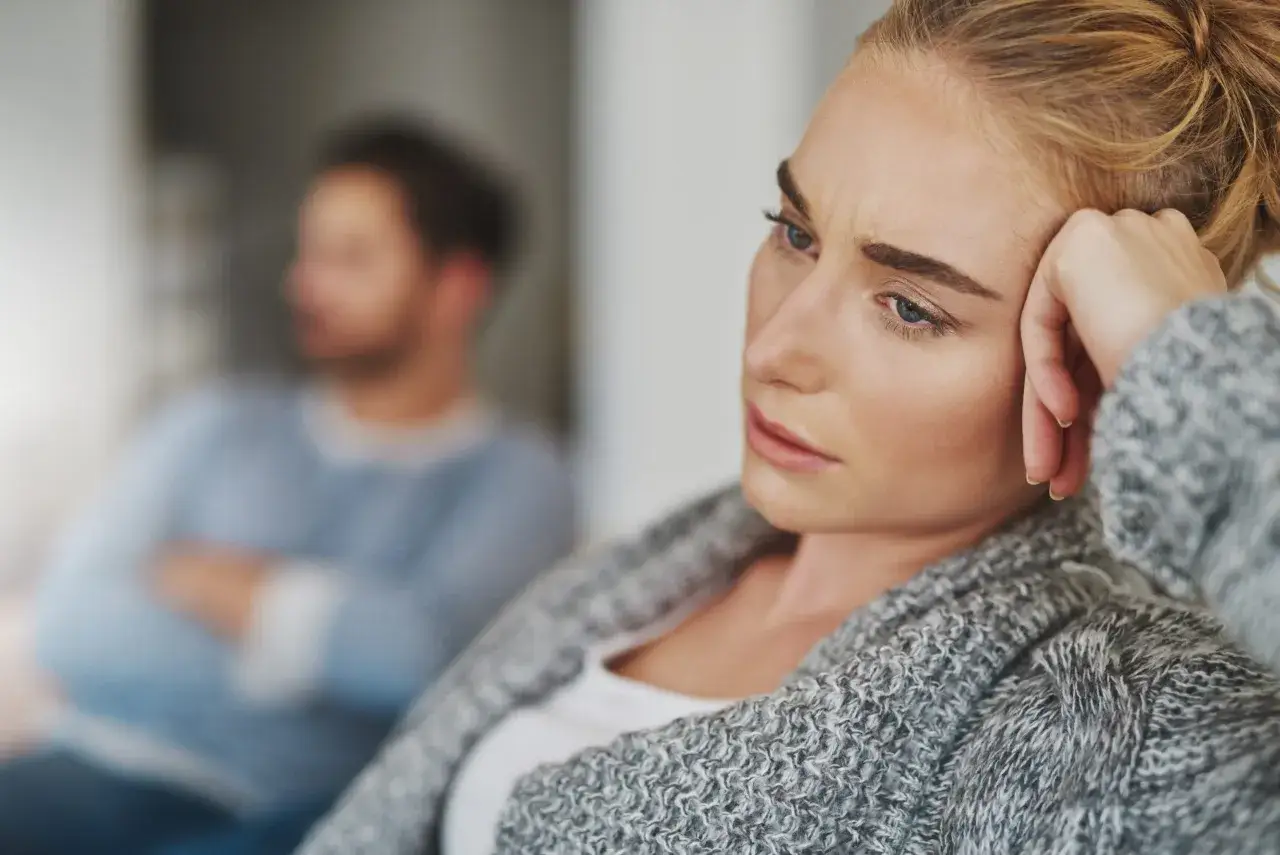 Mujer con expresi&oacute;n de tristeza y un hombre de espaldas, ambos sentados, sugiriendo un matrimonio infeliz y distancia emocional.