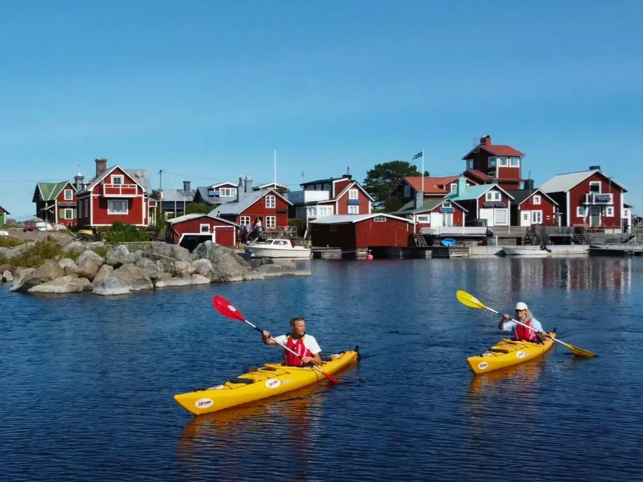 Två personer padlar kajak i en vacker skärgårdsmiljö med röda hus i bakgrunden.