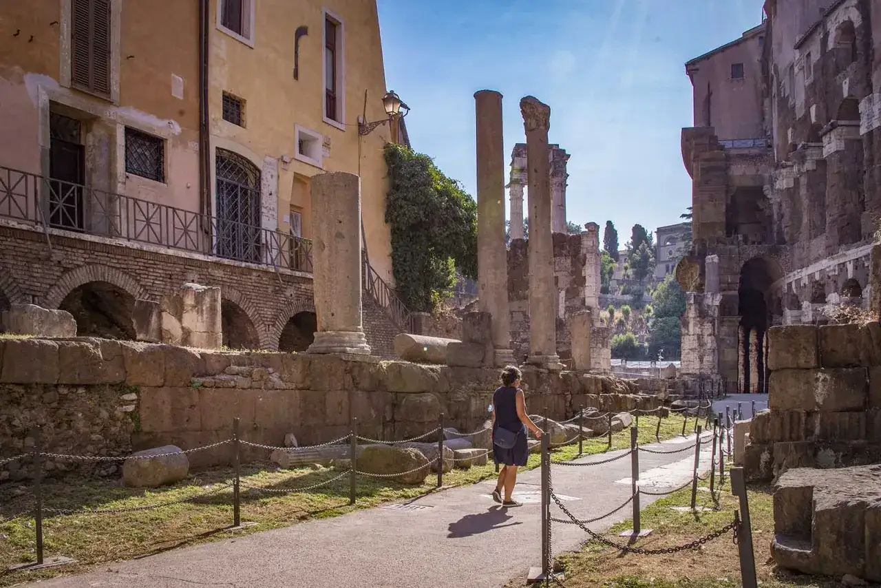 Una donna passeggia tra le rovine del Portico d'Ottavia, con antiche colonne e mura che si stagliano sotto il sole romano.