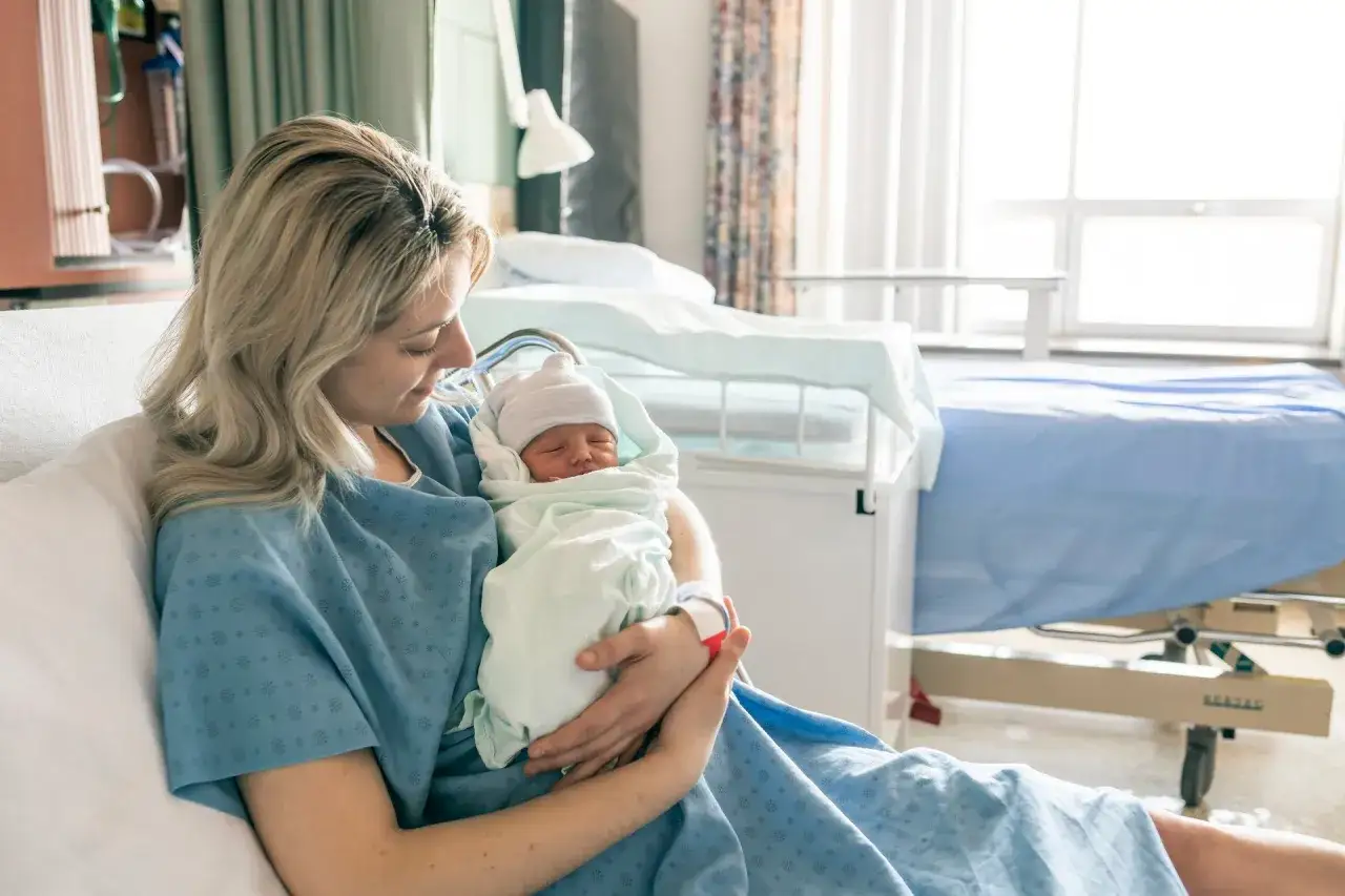 Szczęśliwa mama tuli noworodka. W szpitalnej sali, po porodzie, czeka ją czas regeneracji i opieki nad maleństwem.