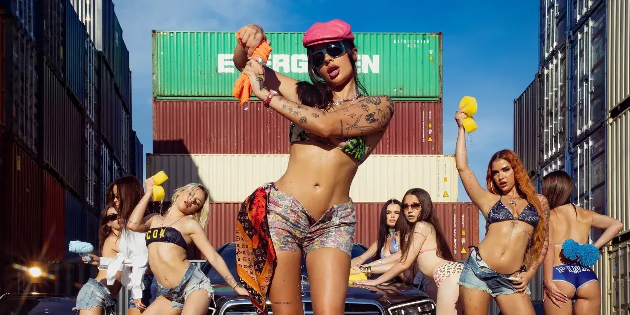 Kobiety w bikini myją auto, trzymając gąbki i szmatki. W tle piętrzą się kontenery, tworząc industrialny klimat dla tych letnich, radosnych fagata utwory.