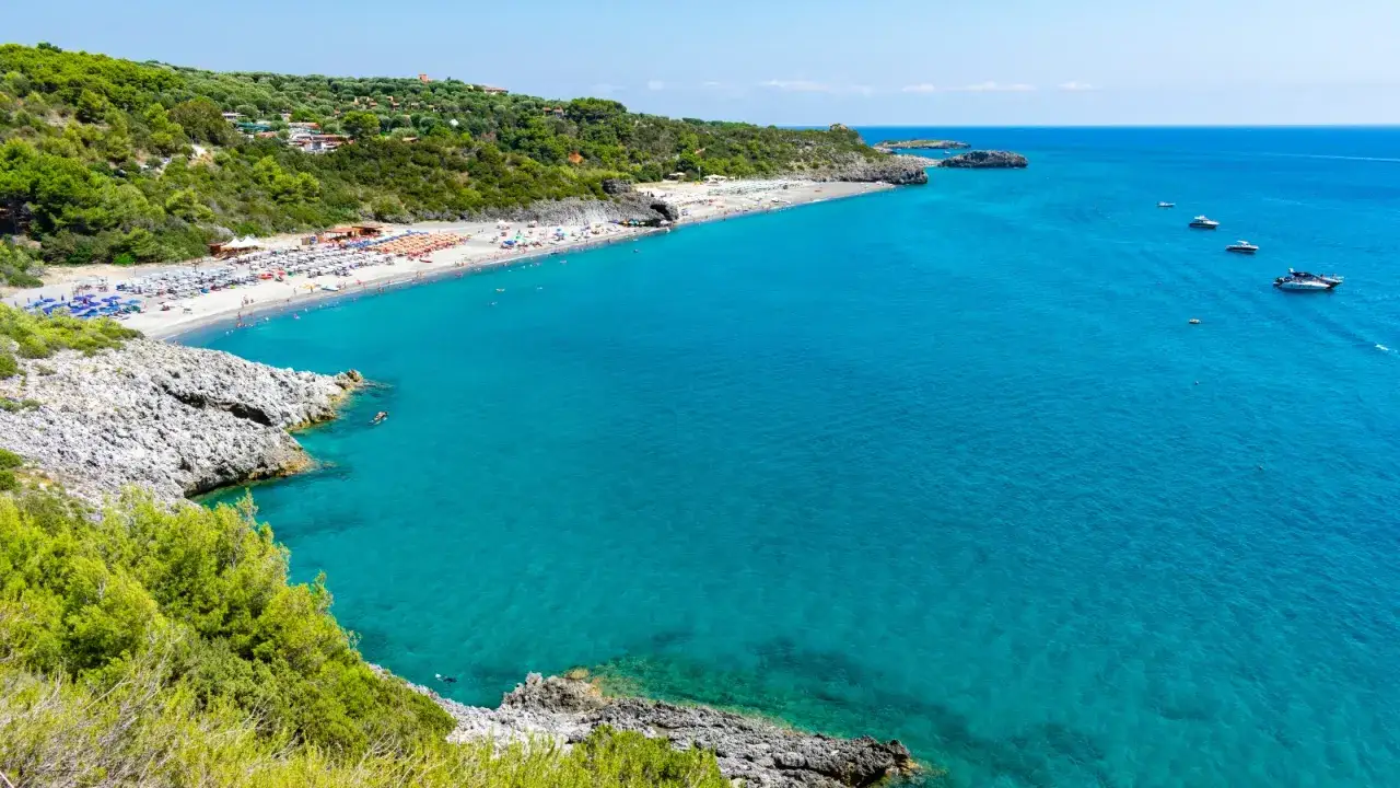 Spiaggia affollata con ombrelloni colorati e mare cristallino nel Cilento. Barche a vela e yacht solcano le acque turchesi.