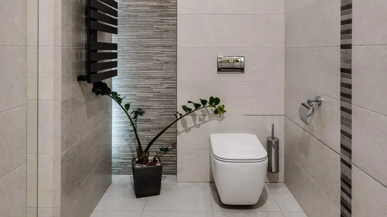 Nowoczesna łazienka z białym WC w rogu, ozdobiona zieloną rośliną i chromowanymi akcesoriami.