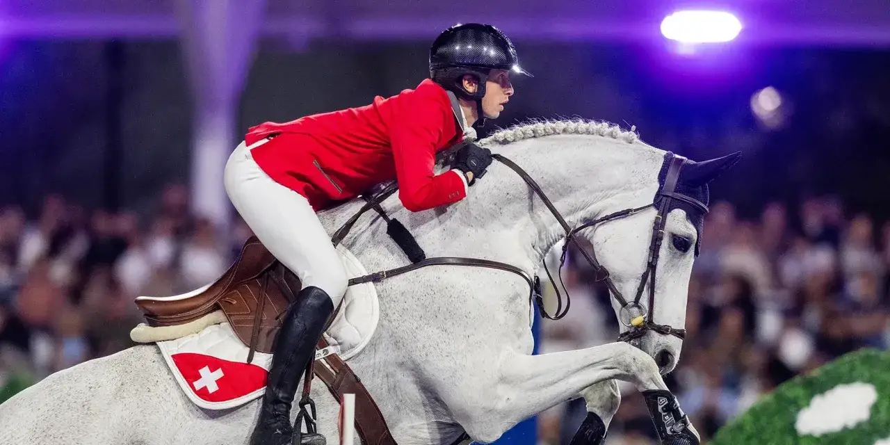 Une cavalière en tenue rouge et blanche, sur un cheval gris, en plein saut. L'équipe de jumping suisse est prête pour la compétition.