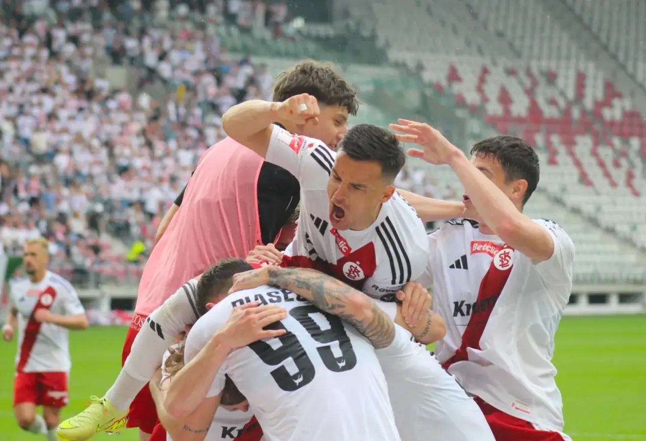 Piłkarze w białych strojach świętują awans do ekstraklasy. Radość i euforia na boisku, trybuny wypełnione kibicami.