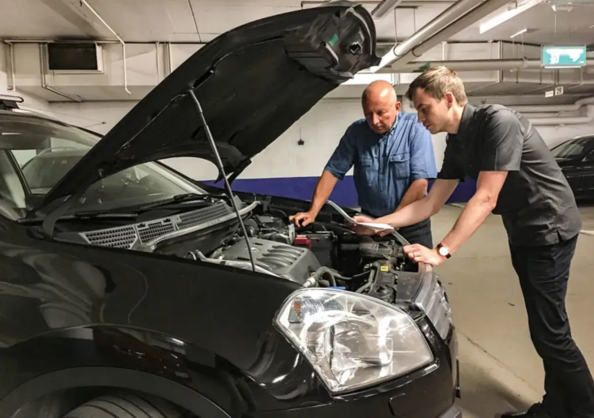 Två män tittar på bilmotorn med motorhuven öppen. En av dem håller i papper, kanske för att se om det är dags att serva bilen varje år.