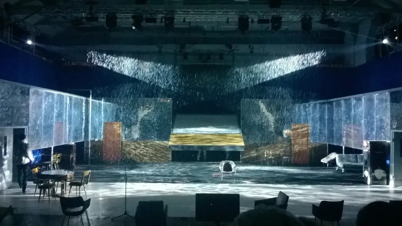 Scena w Teatrze Nowym w Warszawie, z deszczem padającym z góry. W tle nowoczesna scenografia z elementami wody i szkła.