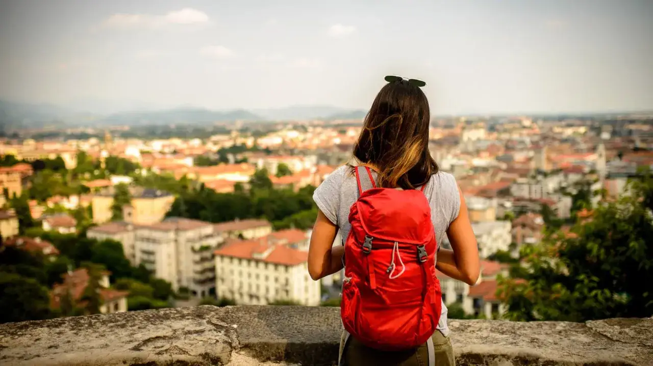 Ragazza con zaino rosso ammira la città dall'alto, pronta per una camminata veloce tra i vicoli.