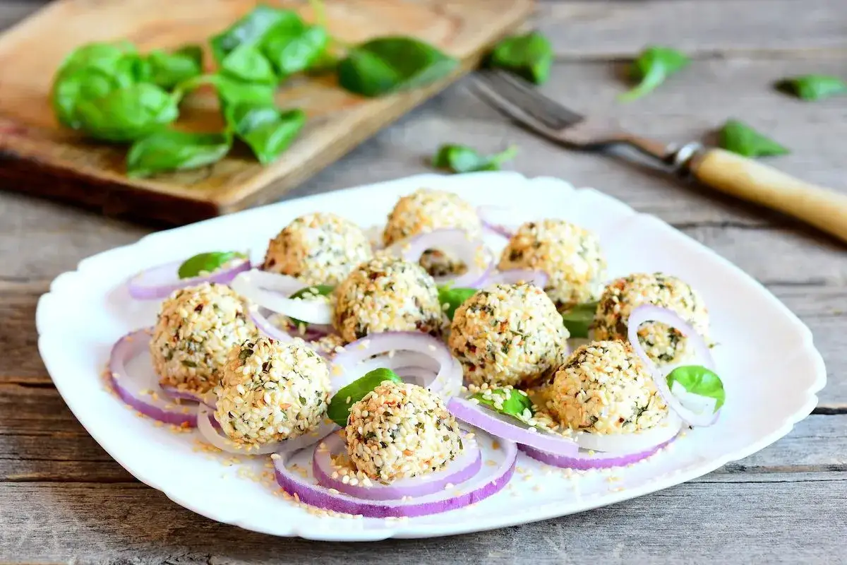 Deliziose polpette di ricotta impanate con semi di sesamo, servite su anelli di cipolla rossa e foglie di basilico fresco.