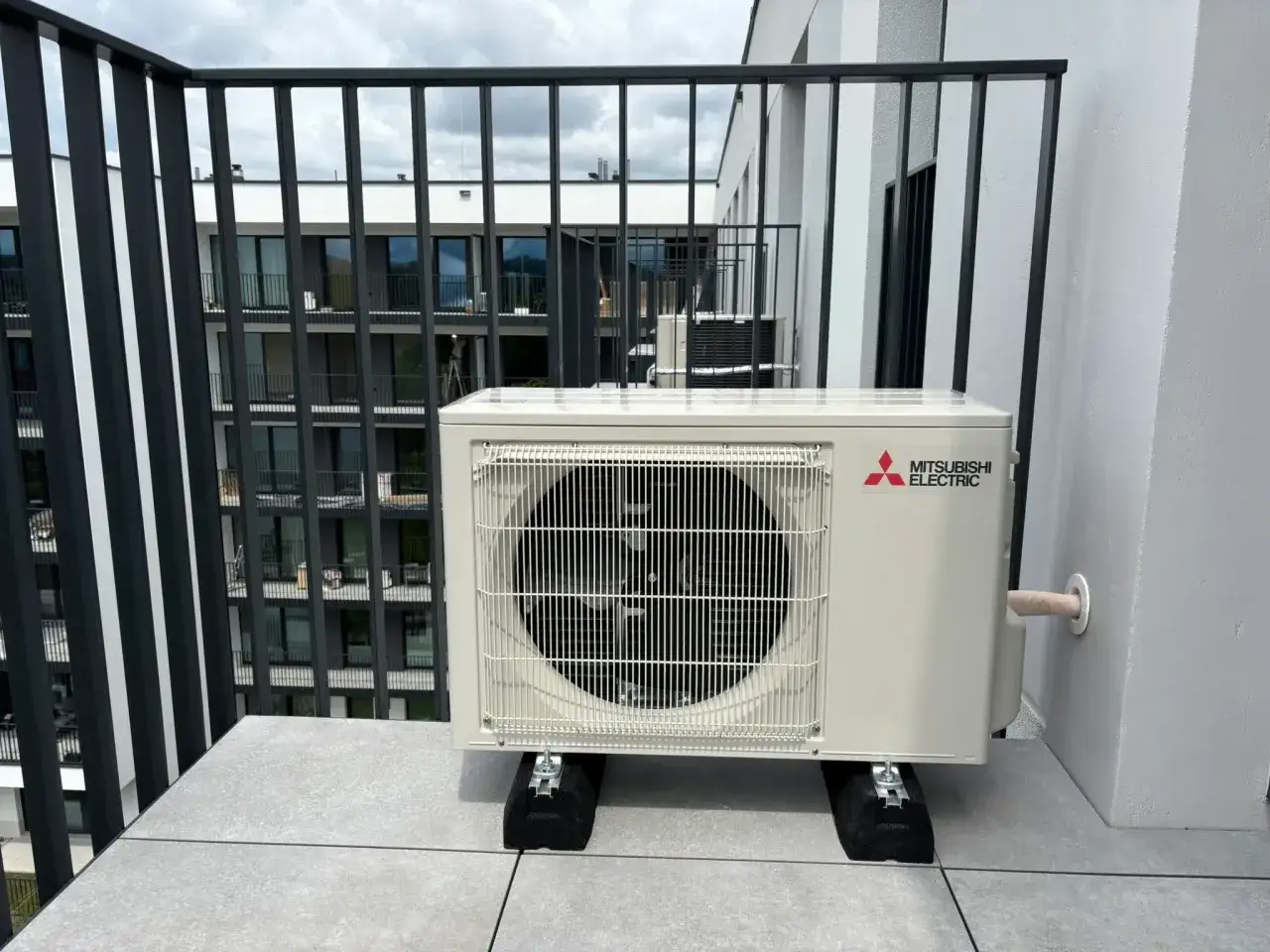 Jednostka zewnętrzna klimatyzatora Mitsubishi Electric na balkonie z widokiem na budynek mieszkalny.