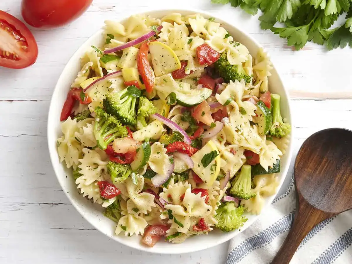 Ein bunter, sommerlicher Nudelsalat mit Farfalle, Brokkoli, Tomaten, Zucchini und roten Zwiebeln, serviert in einer wei&szlig;en Sch&uuml;ssel.