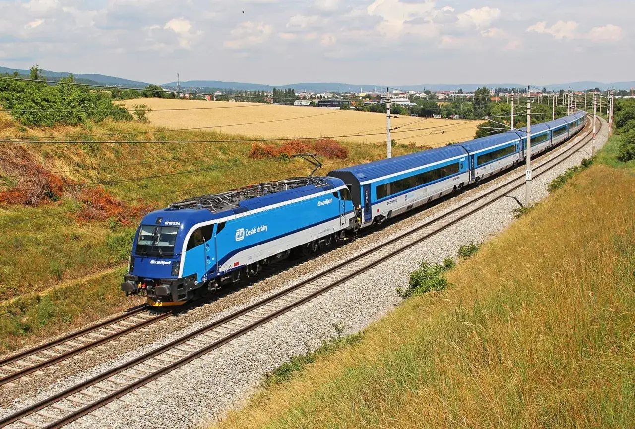 Niebieski pociąg Railjet czeskich kolei mknie po torach przez krajobraz z polami i miastem w tle. Idealny sposób, by tanio do Pragi pociągiem!
