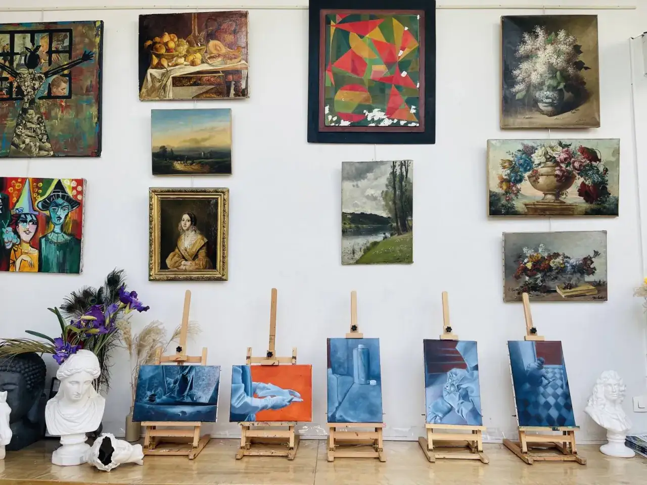 Atelier d'art avec des toiles diverses, dont des natures mortes et des portraits, prêtes à peindre à l'huile. Des chevalets présentent des œuvres en cours.