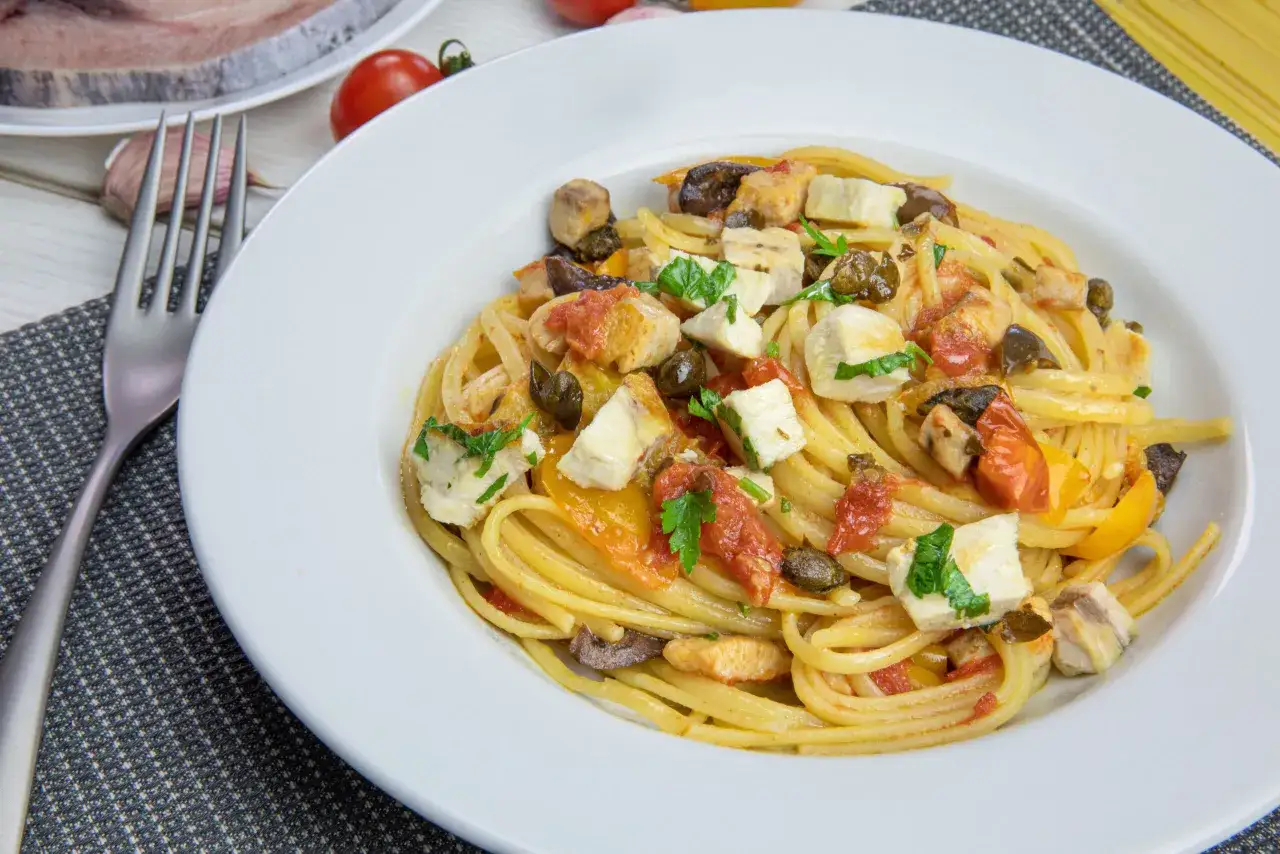 Un piatto di pasta con pesce spada, pomodorini, olive e capperi, guarnito con prezzemolo fresco.
