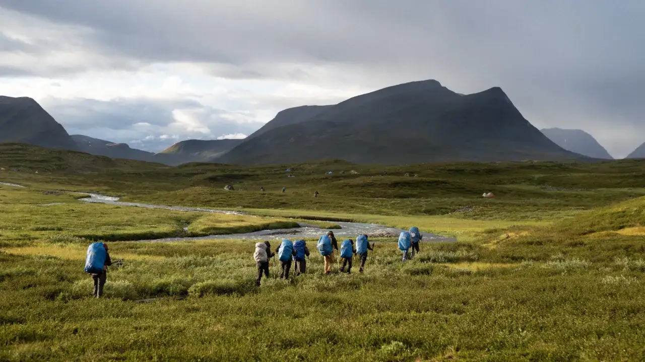 Hikers with large backpacks trek through a grassy valley in Sweden, with mountains in the distance. This is the Fj&auml;llr&auml;ven Classic Sweden.