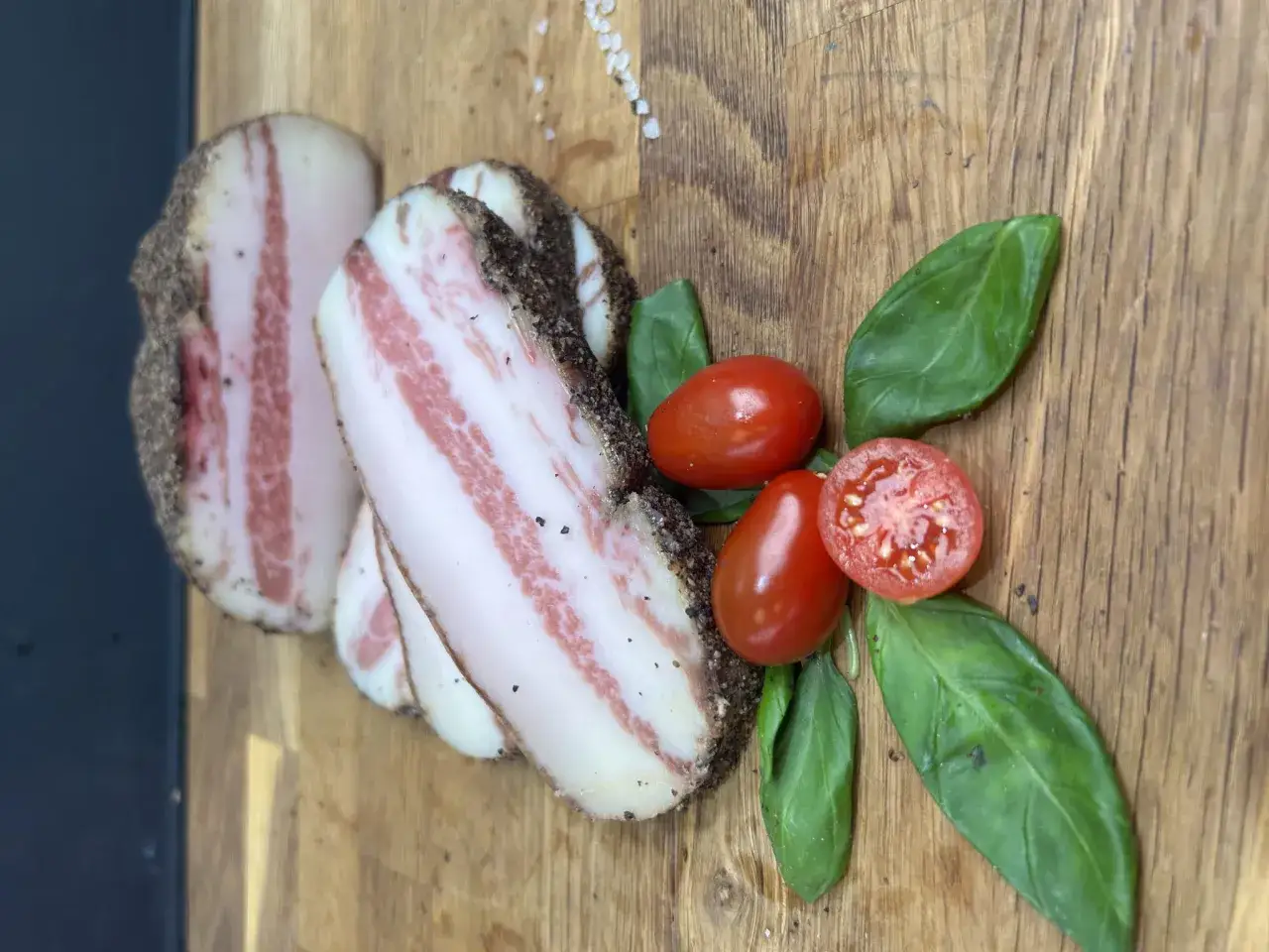 Plastry guanciale, czyli wędzonego podgardla wieprzowego, z pieprzem, pomidorkami koktajlowymi i bazylią na desce.
