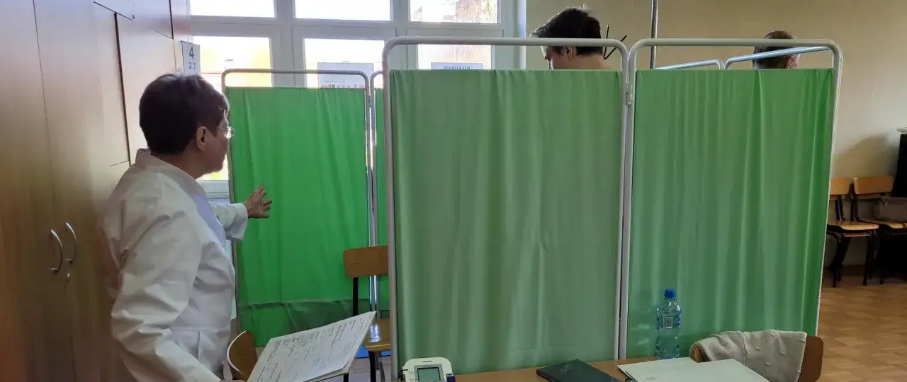 Lekarka w białym kitlu kieruje pacjenta za zieloną zasłonę. To może być gabinet lekarski, ale też punkt badań dla żołnierzy. Kategoria B wojsko.