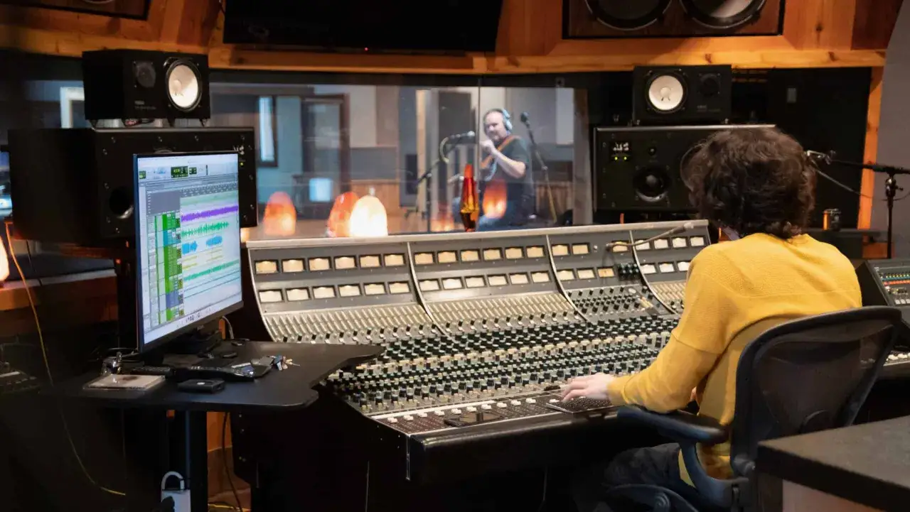 Ingénieur du son ajustant les réglages sur une console pour créer un effet de reverb definition musique, avec un musicien en studio derrière la vitre.