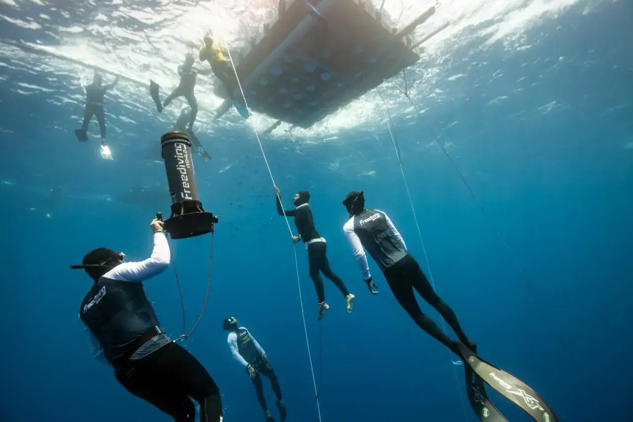 Pod wodą, w błękitnej toni, nurkowie przygotowują się do bicia freediving rekord. Wokół nich widać sprzęt i innych zawodników.